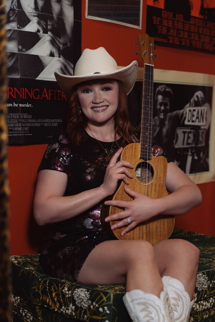 Emily Glover, singer-songwriter, smiling in a cream cowboy hat and holding her ukulele upright.