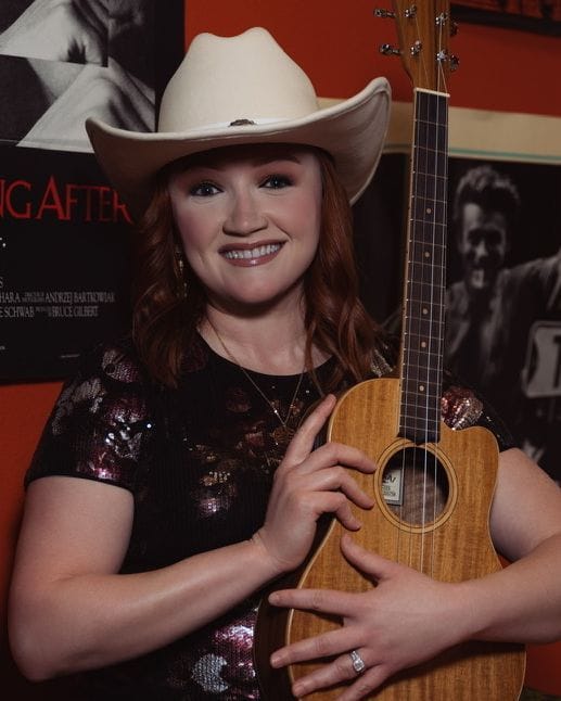 Emily Glover smiling in a cream cowboy hat, holding her ukulele upright.