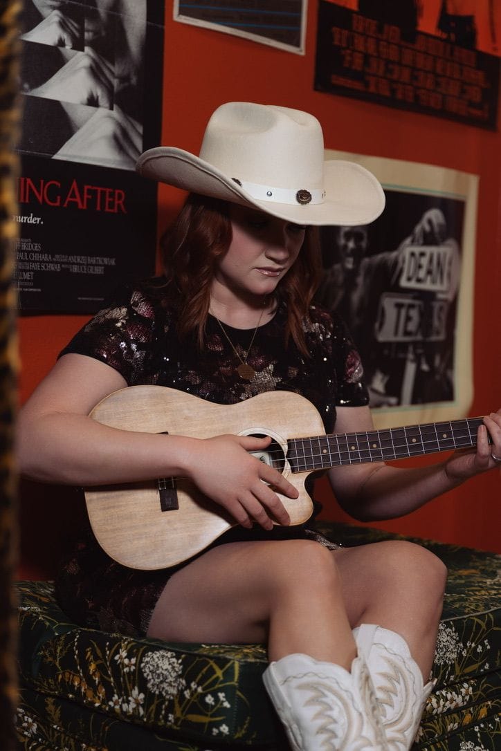 Emily Glover looking down at her ukulele mid-song against a rust-red wall of vintage posters.