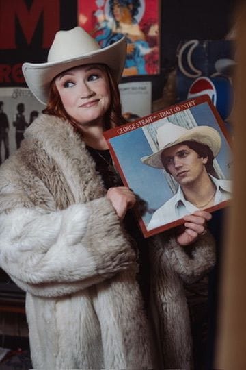 Emily Glover in a fur coat and cowboy hat holding up a vintage country record.