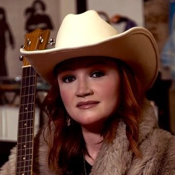 Close portrait of Emily Glover in a cream cowboy hat and fur-trimmed coat with a ukulele headstock beside her.